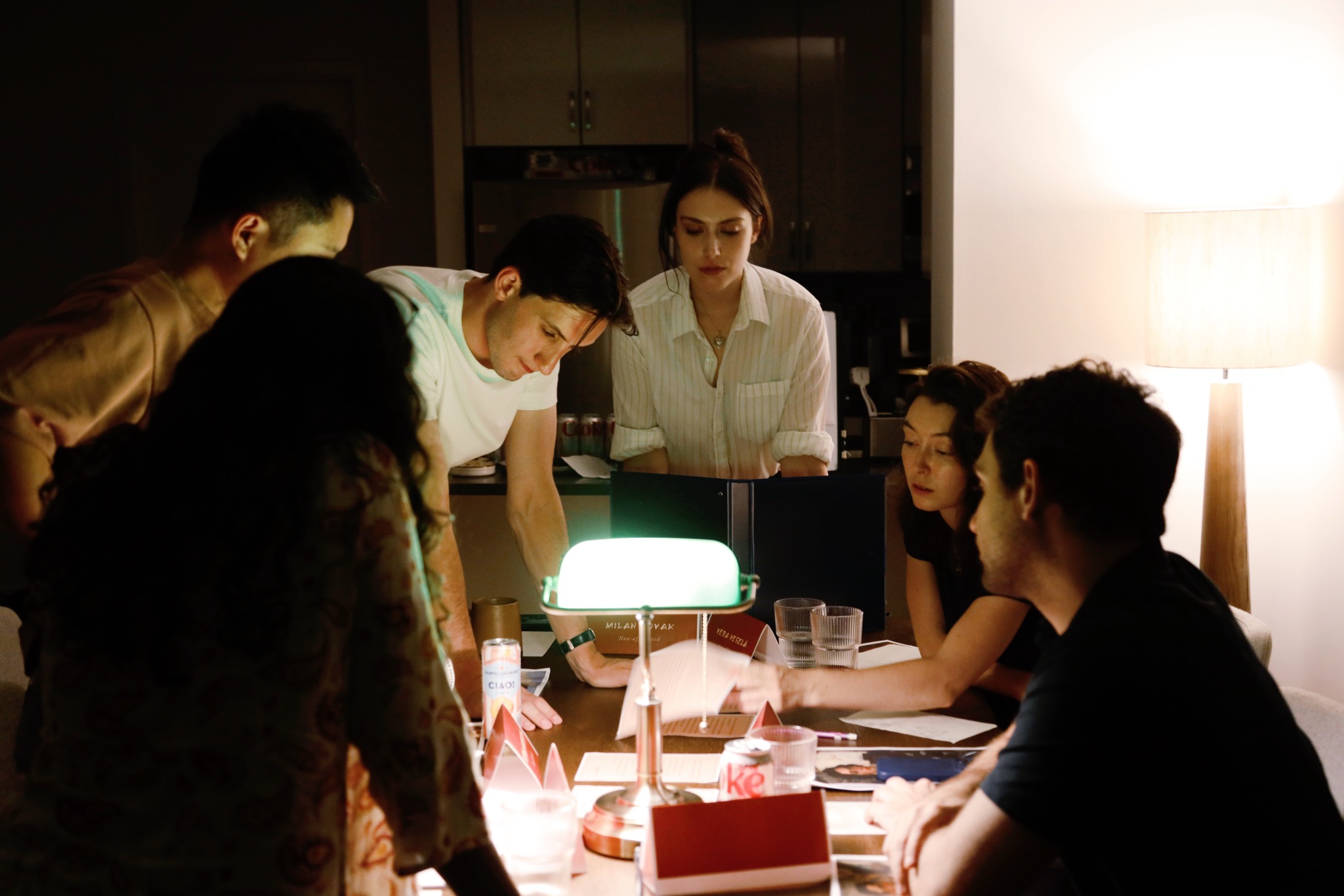 A cast of six gathered around a lamplit table in character